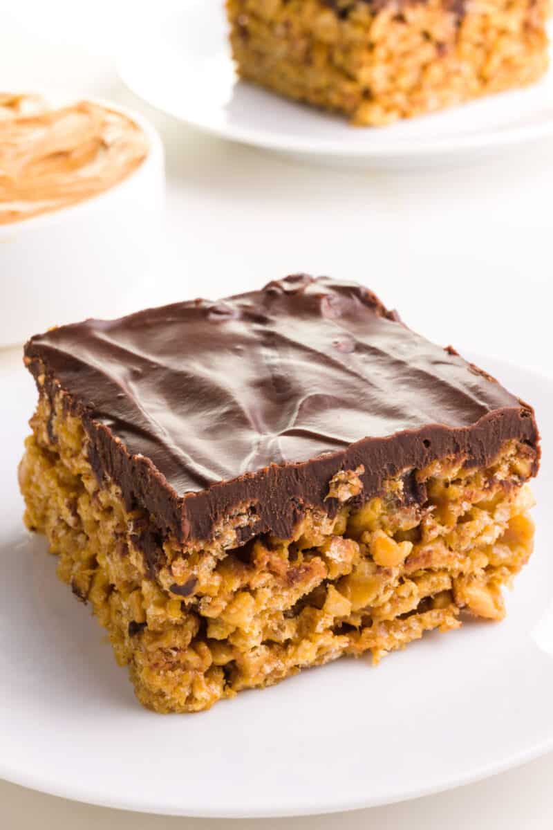 A vegan special K bar sits on a plate in front of a bowl of peanut butter and another treat in the background.